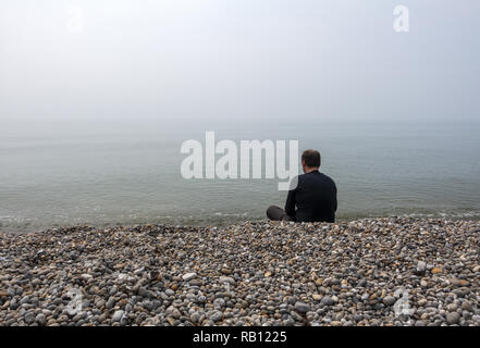 Uomo seduto da solo sulla spiaggia di ciottoli che si affaccia sul mare coperto di nebbia Foto Stock