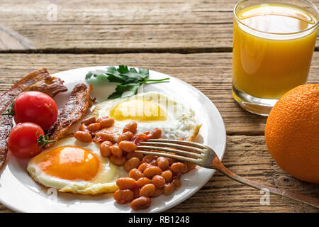 Inglese Colazione La colazione è servita in una piastra con un uovo fritto, fagioli, pomodori e pancetta con succo d'arancia fresco su un tavolo di legno. close up Foto Stock