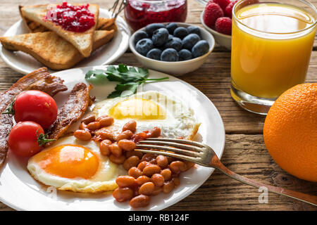 Colazione con succo d'arancia, uova fritte, pancetta, fagioli, frutta e bacche. close up Foto Stock