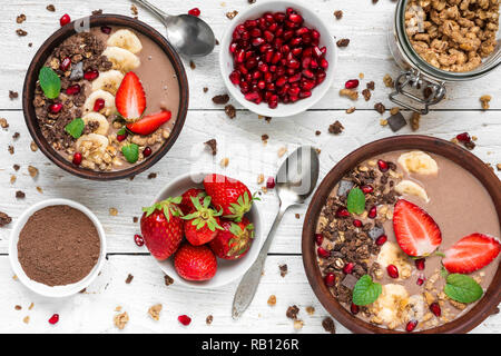 Cacao o cioccolato e banana frullato di proteina ciotole con muesli, fragola e i semi di melograno è servito per la prima colazione con cucchiai in legno bianco t Foto Stock