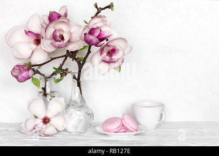 Rosa magnolia bouquet di fiori con tazza di caffè e macarons su bianco sullo sfondo di legno. vacanze ancora vita Foto Stock