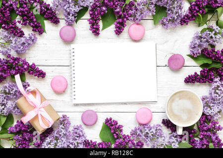 Cornice fatta di fiori lilla con bigliettino vuoto o invito a nozze con tazza di caffè, confezione regalo e macarons su bianco tavolo in legno. mock up. fl Foto Stock