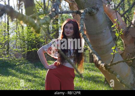 Una giovane donna con lunghi brunette capelli e vestiti alla moda in rosso pantaloni svasati appoggia contro un argento betulla in un bosco inglese in primavera Foto Stock