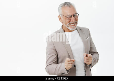 Unretouched studio shot di elegante e felice carismatico imprenditore maturo in bicchieri e tuta con cattivo denti winking con gioia il piacere di puntamento a telecamera come se hinting contro il muro grigio Foto Stock