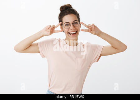 Carismatico e giocoso emotiva di sorella in bicchieri arrotondati con un ampio sorriso che mostra segni di pace vicino a faccia come divertirsi giocando e ingannare intorno con i fratelli e le sorelle oltre il muro grigio Foto Stock