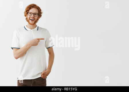 Felice entusiasta redhead bello ragazzo con barba lunga in bicchieri e abbigliamento casual rivolta a destra con il dito indice e sorridente largamente mentre viene sollevato e suggerendo amici rimanere al suo posto Foto Stock