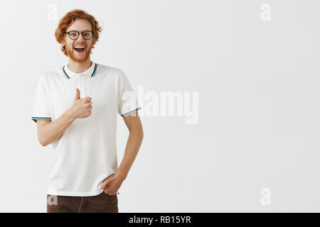 Studio shot di sostegno hanno intrattenuto e divertito felice redhead barbuto ragazzo nel casual Polo shirt e occhiali neri sorridente largamente mostrando pollice in alto gradimento idea o il tifo qualcuno per bene allo sforzo Foto Stock