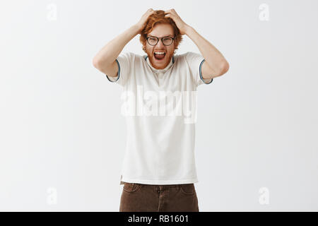 Non è in grado di trattenere le emozioni negative più interno alimentato di pressione. Ritratto di med e insane urlando redhead uomo con barba tira i capelli al di fuori della testa urla con ira e con sdegno su sfondo grigio Foto Stock