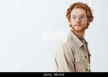 Studio shot di sognante bel giovane redhead guy passando da interessanti store e girando a sinistra con calma espressione essendo portati via con pensieri indossando giacca beige e vetri trasparenti Foto Stock