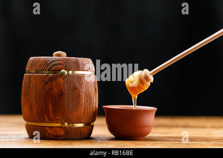 Canna con miele e un cucchiaio di pettinatura a pettine fuori il miele su una tavola di legno contro uno sfondo scuro. Close-up. Foto Stock