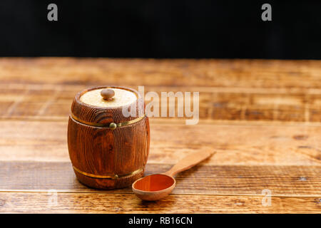 Chiuso il barile con il miele e un cucchiaio di legno su una tavola di legno su uno sfondo scuro. Canna. Close-up. Foto Stock