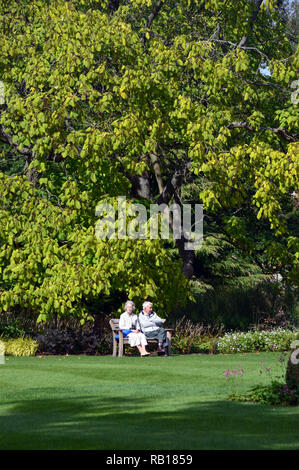 Coppia Matura seduta su una panca in legno su un prato sotto il sole a RHS Garden Harlow Carr, Harrogate, Yorkshire. Inghilterra, Regno Unito. Foto Stock