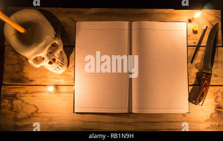 Cranio umano con la luce di una candela e notebook con una matita e il coltello sul tavolo in legno e decorate per tema Halloween con copia spazio. Foto Stock