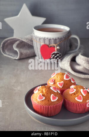 Muffin decorati con cuori di zucchero e una tazza con cuore rosso su sfondo grigio chiaro. Il giorno di San Valentino o in inverno il concetto di compleanno. Foto Stock