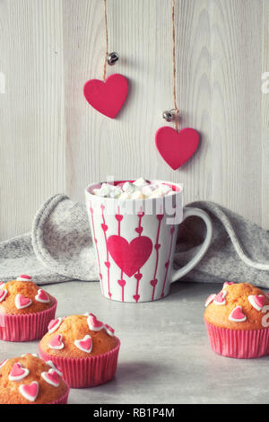 Muffin decorati con cuori di zucchero e una tazza con cuore rosso su sfondo grigio chiaro. Il giorno di San Valentino o in inverno il concetto di compleanno. Foto Stock