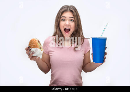 Felice giovane donna urlando sulla fotocamera. Ella tenere burger e bicchiere di coca cola in mani. Isolato su sfondo bianco. Foto Stock
