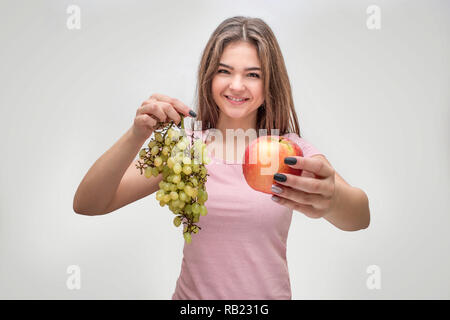 Allegro giovane donna stand e sorriso sulla fotocamera. Ella tenere uve e Apple. Isolato su sfondo grigio. Foto Stock