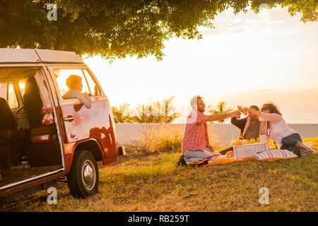 La famiglia e gli amici tutti insieme in picnic di attività per il tempo libero su un prato con un vecchio rosso vintage van parcheggiato e figlio i bambini all'interno di li loooking clinkin Foto Stock