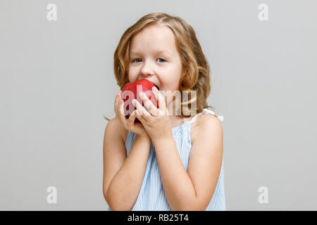 Ritratto di un sorridenti bimba bionda su sfondo grigio. Baby i morsi di mela rossa Foto Stock