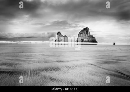 Due rocce e due escursionisti sulla spiaggia Wharariki su nuvoloso giorno di moody a pressione atmosferica immagine in bianco e nero Foto Stock