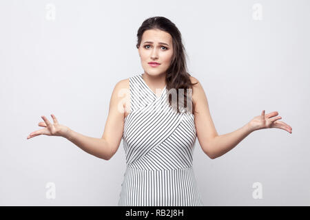 Ritratto di confuso bella bruna giovane donna con il trucco e abito a strisce in piedi e guardando la fotocamera con bracci incrociati e chiedendo a. interna Foto Stock