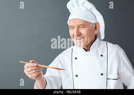 Senior chef maschio studio isolato in piedi sul muro grigio tenendo una spatola di legno piatto degustazione sorridente gioioso close-up Foto Stock