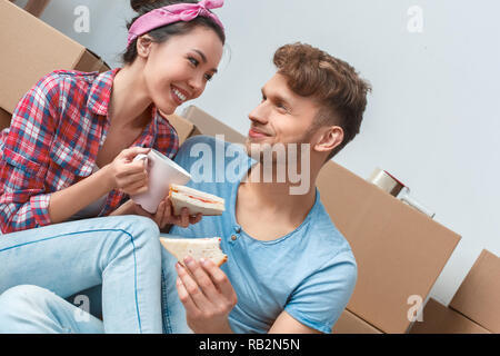 Giovane marito e moglie spostando di nuovo posto seduto sul pavimento con la colazione mangiando panini di bere il caffè caldo guardando ogni altra sorridendo felice Foto Stock