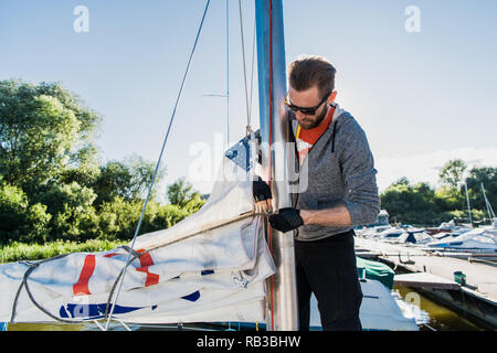 Giovane marinaio in occhiali da sole trattiene e sposta vela principale con entrambe le mani. Egli è felice e sorridente. Giovane uomo è la preparazione di yacht a vela. C'è il sole all'esterno. Foto Stock