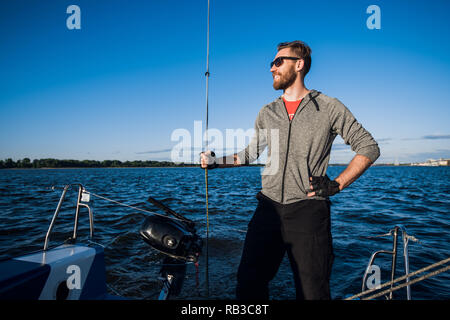 Giovane uomo che indossa gli occhiali da sole in piedi sulla poppa di yacht e godendo di una perfetta giornata autunnale sotto le vele - vacanze a vela nozione Foto Stock