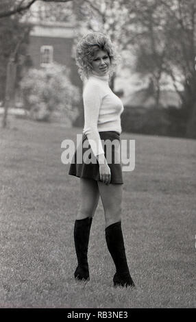 Anni sessanta, sinuosa donna con grandi capelli ondulati e indossa una mini gonna, tartaruga-maglione collo e stivali in pelle che pongono al di fuori, Inghilterra, Regno Unito. Foto Stock