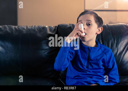 8 anno vecchio ragazzo di indossare abbigliamento sportivo inalazione con un inalatore per asmatici seduto in divano. I concetti : salute, malattia, il doping Foto Stock