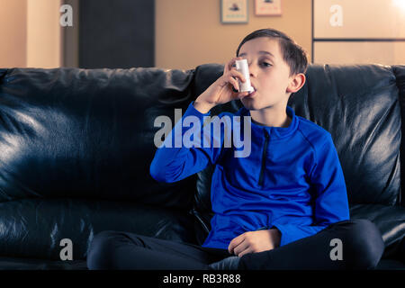 8 anno vecchio ragazzo di indossare abbigliamento sportivo inalazione con un inalatore per asmatici seduto in divano. I concetti : salute, malattia, il doping Foto Stock