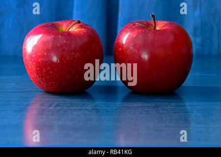 Due brillanti mele rosse su blu riflettente piano tavolo e sfondo blu, laterale vista da vicino Foto Stock