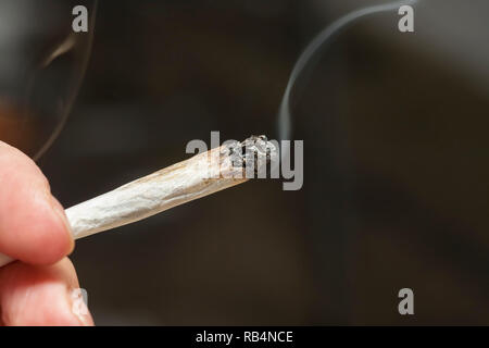 Close-up immagine di una mano che tiene un acceso marijuana joint contro sfondo grigio scuro Foto Stock
