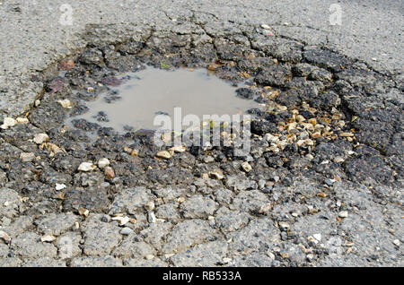 Un perfido buca in una strada asfaltata. Pieno di acqua a pietre sciolte, questa buca è stato creato durante il cattivo tempo invernale. Foto Stock