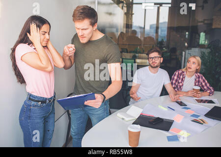 Angry Young amn fissare plastica compressa merita di carte e il punto su di esso. Si è arrabbiato e urlando. Altre due persone sedersi dietro di loro. Essi sono rimasti stupiti e spaventati. Brunette tenere le orecchie e gli occhi chiusi. Foto Stock