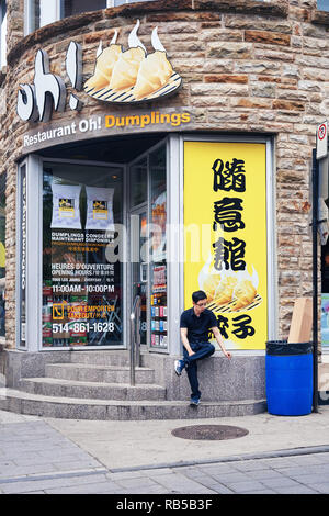 Giovane maschio asiatico seduto su una parete di un cinese gnocco store fuma sigaretta sulla strada a Montreal, Quebec, Canada. Foto Stock