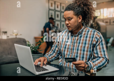 I giovani alla moda di razza mista guy effettuare un acquisto online utilizzando la sua carta di credito in un cafe Foto Stock