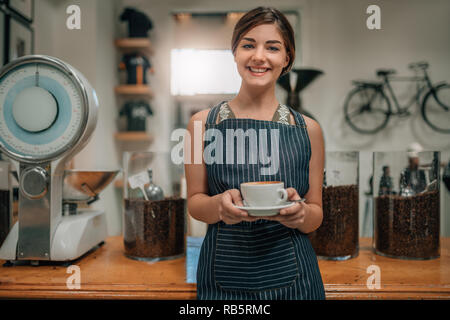 Giovane bella cameriera sorridente che serve caffè con latte art permanente, mentre in coffee shop. Foto Stock