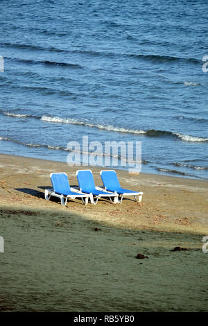 Bassa stagione. Vuoto spiaggia sabbiosa con tre in plastica bianca lettini per prendere il sole sulla sabbia vicino prima seacoast bordo mare con piccole onde sulla luminosa giornata di sole Foto Stock