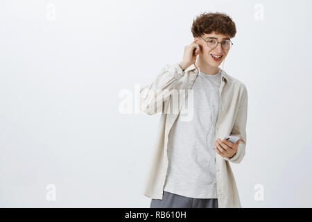 Tiro al coperto del carismatico felice e spensierato giovane europeo modello maschile con capelli ricci in bicchieri arrotondati mettendo sul cuscinetto come l'ascolto di musica con app per smartphone a sorridere soddisfatto della fotocamera Foto Stock