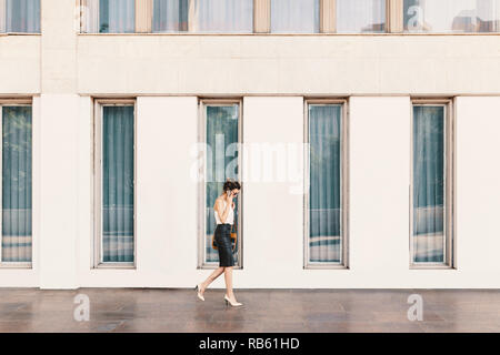 La squisita business donna in pelle gonna e camicia di seta a parlare al telefono mentre si cammina nei pressi di un edificio con grandi finestre. Business, comunicazione Foto Stock