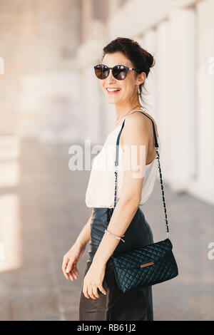 Bella sorridente giovane donna in occhiali da sole che indossa la frizione in pelle e mantello bianco facile camicetta e guardando lontano sopra la spalla mentre si cammina superarvi Foto Stock