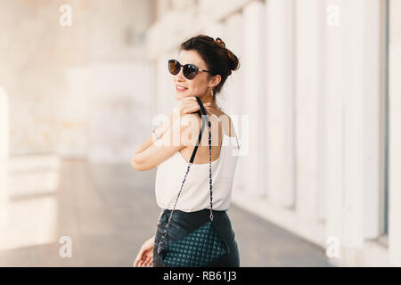 Bella sorridente giovane donna in occhiali da sole che indossa la frizione in pelle e mantello bianco facile camicetta e guardando lontano sopra la spalla mentre si cammina superarvi Foto Stock