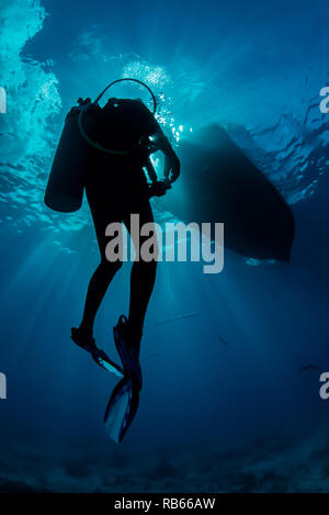 Diver al suo modo di superficie con raggi di sole piercing attraverso la superficie di acqua con il mare azzurro acqua e silhouette in barca sulla superficie. Foto Stock