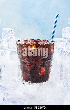 Estate bere caffè ghiacciato o soda in un bicchiere con goccioline e paglia circondati da cubetti di ghiaccio sulla neve su sfondo blu. Messa a fuoco selettiva, spazio di copia Foto Stock