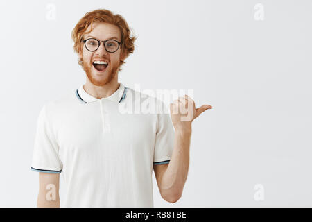 Emozionato overemotive funny redhead modello maschile con barba disordinato nel polo shirt e bicchieri rivolta a destra con il pollice e sorridente retelling gioiosamente interessante e una straordinaria storia di successo in questo modo Foto Stock