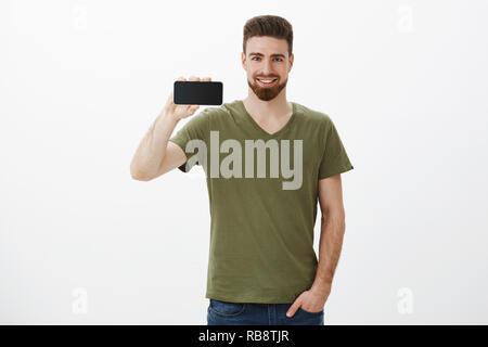 Check out questa incredibile display. Ritratto di fiducioso bello barbuto collega maschio di contenimento orizzontale dello smartphone e sorridendo felice come se vantarsi circa le nuove app o dispositivo, promozione di telefono mobile Foto Stock