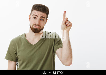 Guy hanno un suggerimento sollevando il dito indice aggiungere idea sciocca permanente e carino con esitanti e timido broncio espressione testa tiltante e guardando la telecamera con sorriso carino, rivolto verso l'alto oltre il muro bianco Foto Stock