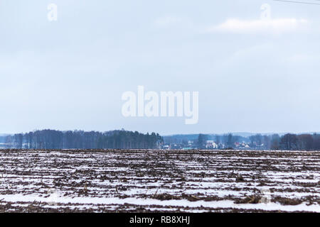 Un po' di neve sul campo arato. Villaggio e la foresta in background. L'inizio dell'inverno in Europa. Foto Stock